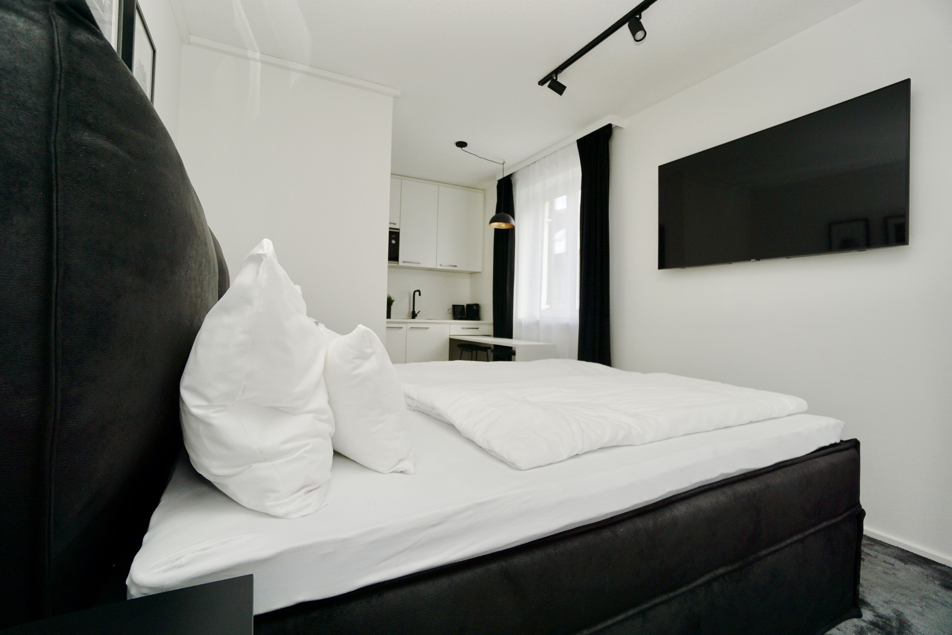 A modern hotel room with a large bed, a flat-screen TV mounted on the wall, and a visible kitchen area in the background.