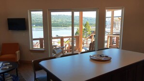 A room with a large window overlooking a lake, a dining table, a TV mounted on the wall, and a chair. - Alderwood Cabin - Homer Seaplane Base (Homer)