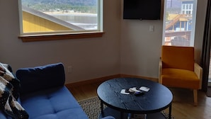 A room with a large window, a flat-screen TV, a blue sofa, a round table, and an orange chair. - Alderwood Cabin - Homer Seaplane Base (Homer)