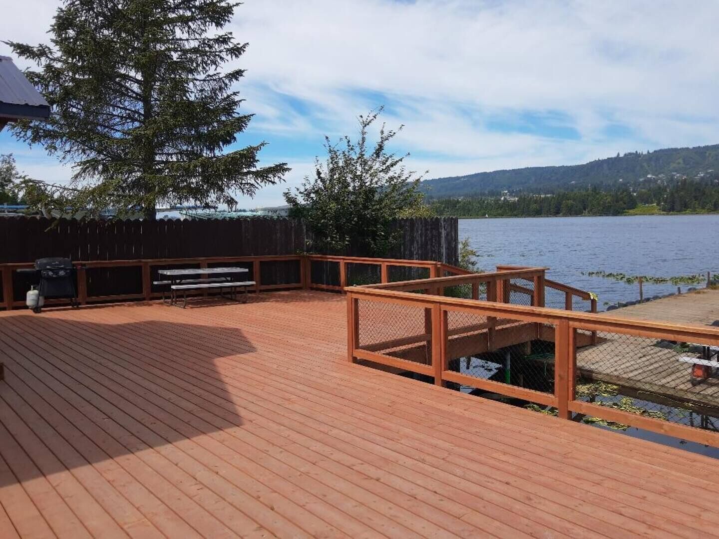 A wooden deck with a grill and picnic table, overlooking a lake with hills in the background.