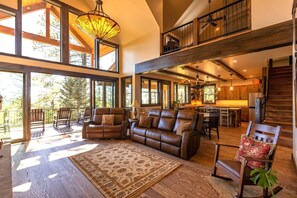 A spacious living area with a large leather sofa, wooden chairs, and a patterned rug. There is a balcony with chairs and a view of trees. - AC, Close to Wolf Creek, Quiet! (Pagosa Springs)