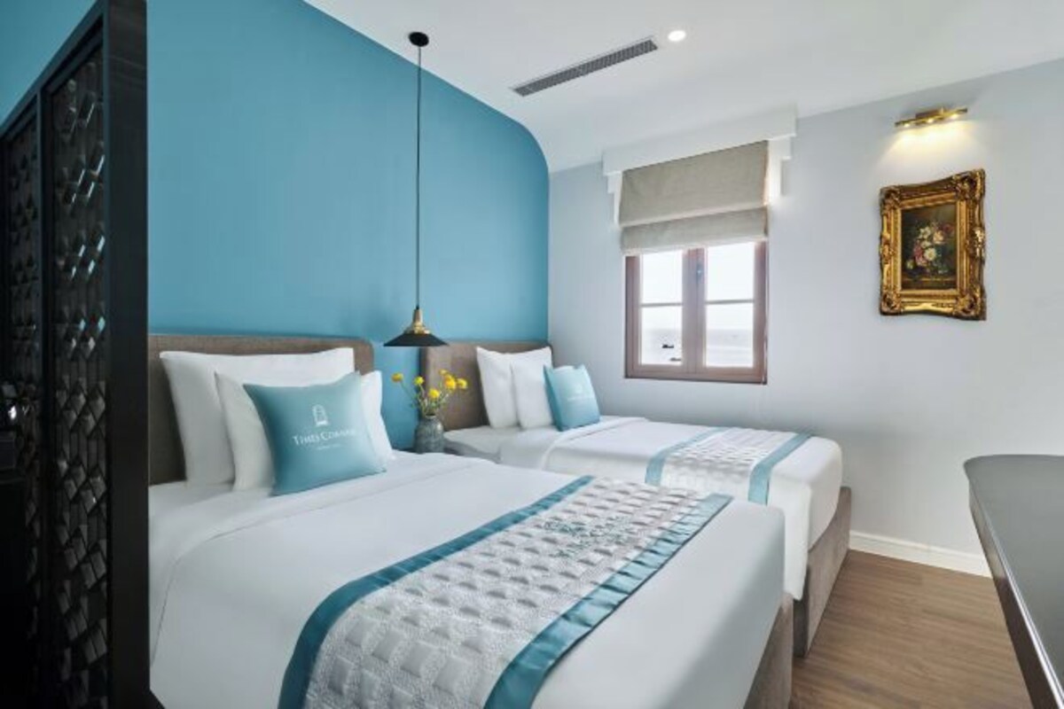 A hotel room with two beds, a window with a view, a painting on the wall, and a ceiling light.