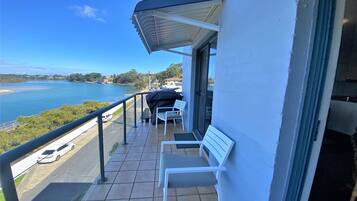 A balcony with a view of a body of water, a covered barbecue grill, and two white chairs.