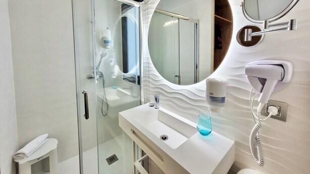 Une salle de bain moderne avec une douche à parois de verre, un lavabo blanc avec un récipient en verre, un sèche-cheveux et un miroir.