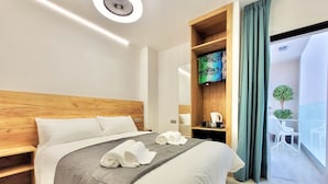 Quarto de hotel moderno com cabeceira de madeira, cama grande com roupa de cama branca, mesa de cabeceira com cafeteira e vista para a varanda.