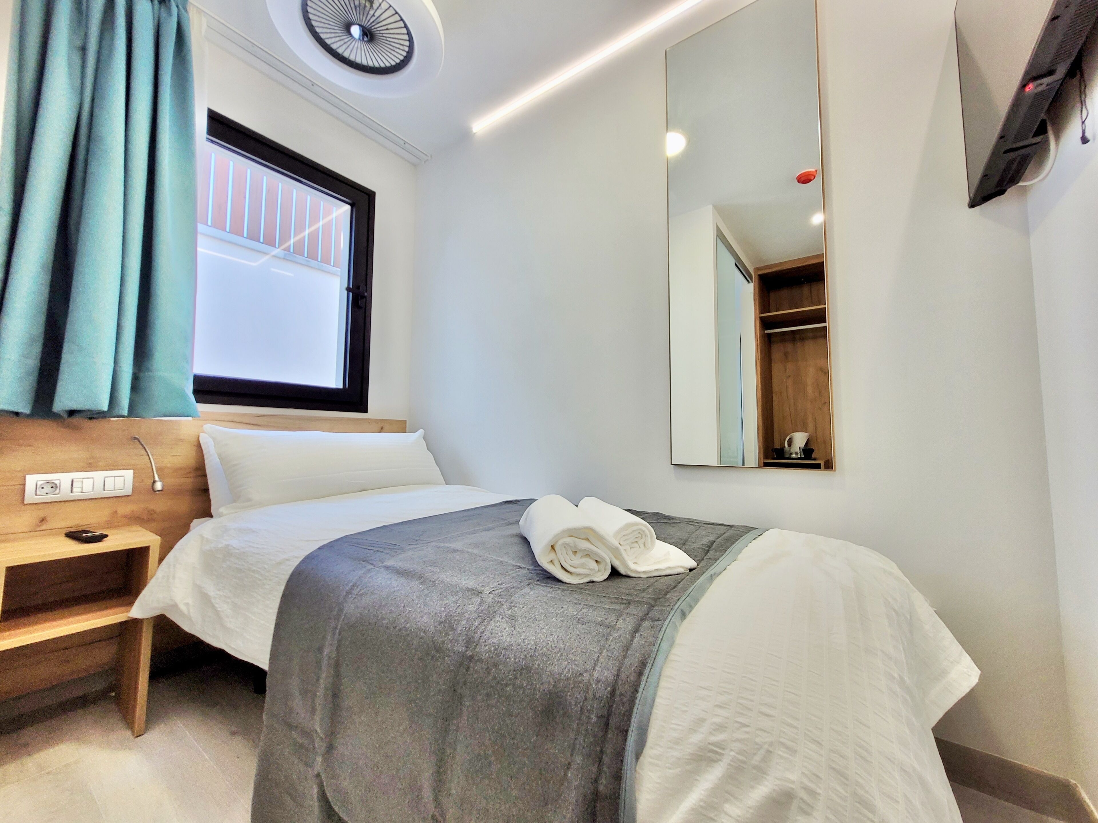 A hotel room with a bed, a bedside table, a mirror, a window with curtains, and a ceiling fan.