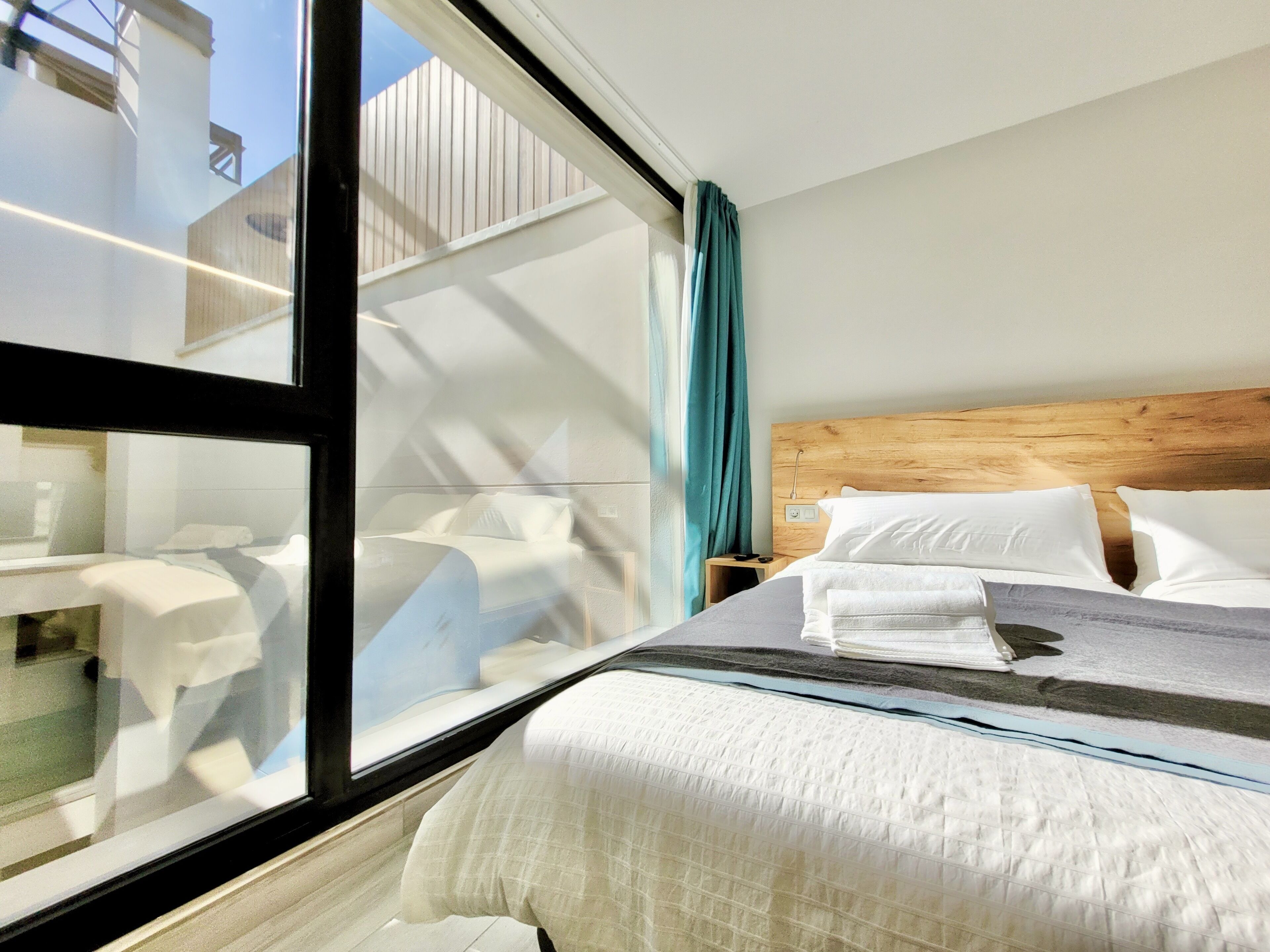 A hotel room with a large window, a wooden headboard, a neatly made bed with white linens, and a small bedside table with a lamp.