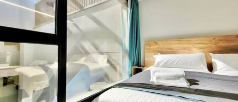 A hotel room with a large window, a wooden headboard, a neatly made bed with white linens, and a small bedside table with a lamp.
