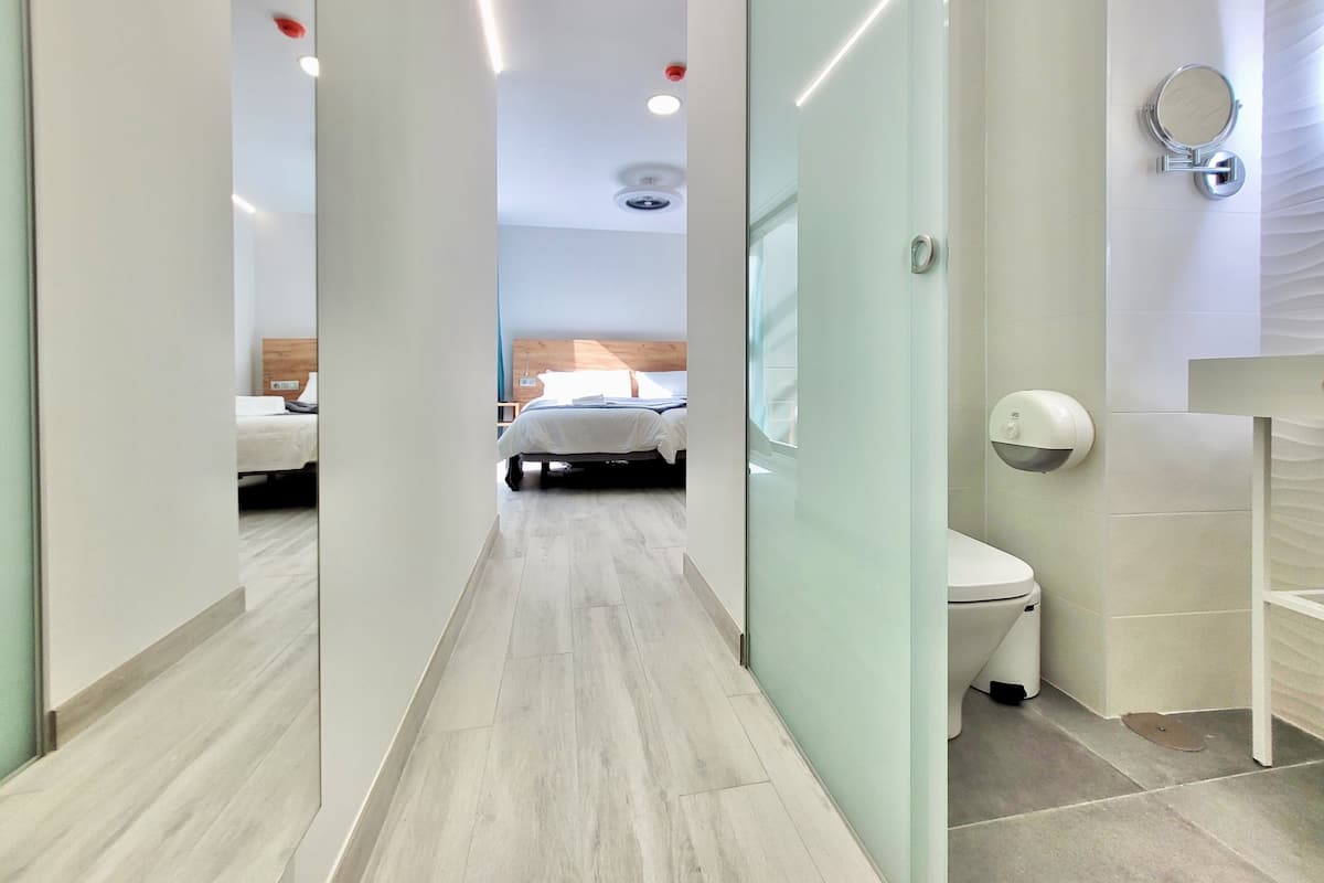 Une chambre d’hôtel moderne, équipée d’un lit, d’une salle de bain avec des toilettes et un sèche-mains, et d’un couloir menant à une autre chambre.
