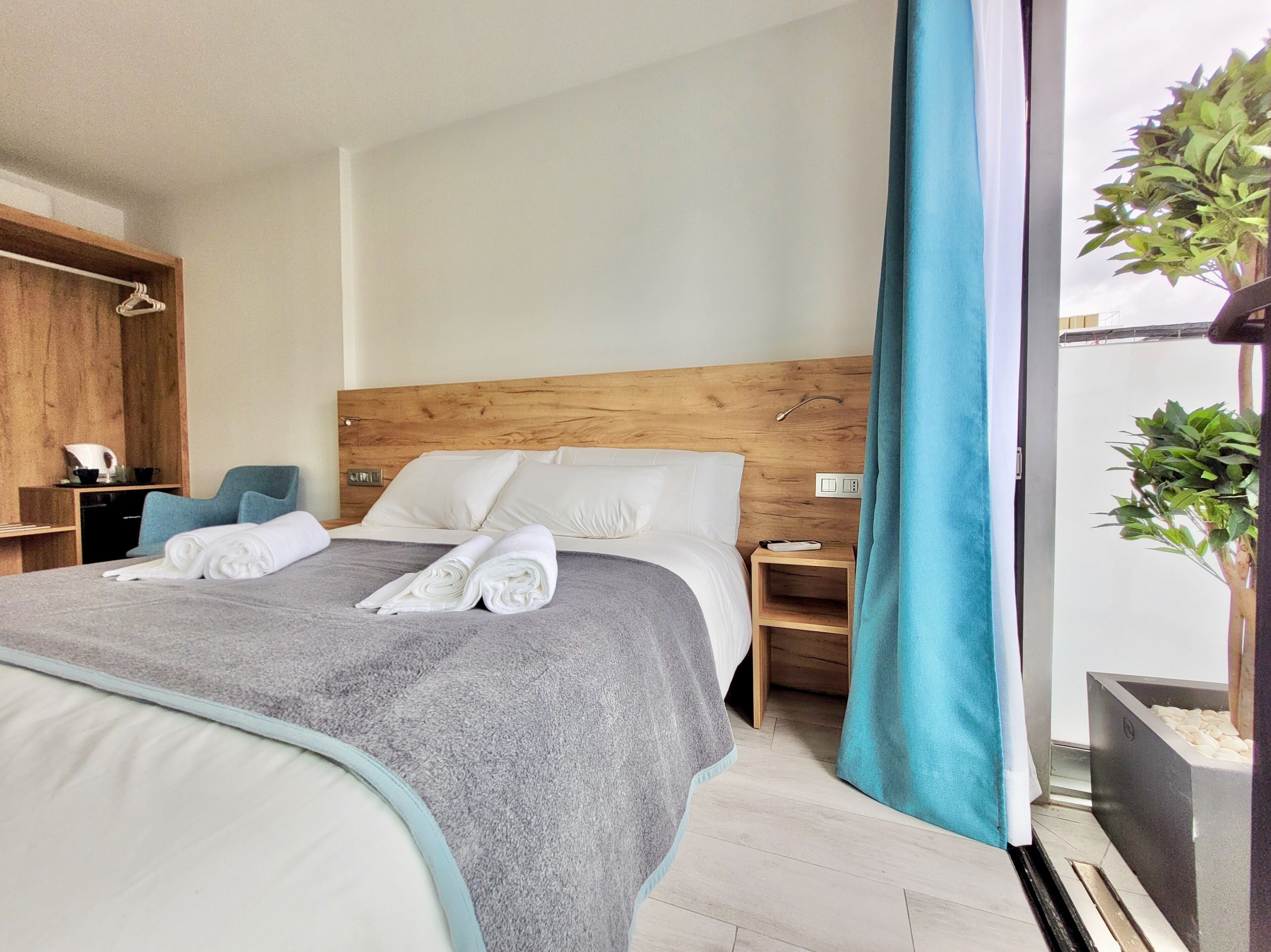 A neatly made bed with white linens and a grey blanket, a wooden headboard, a bedside table with a lamp, and a view of greenery through a window.