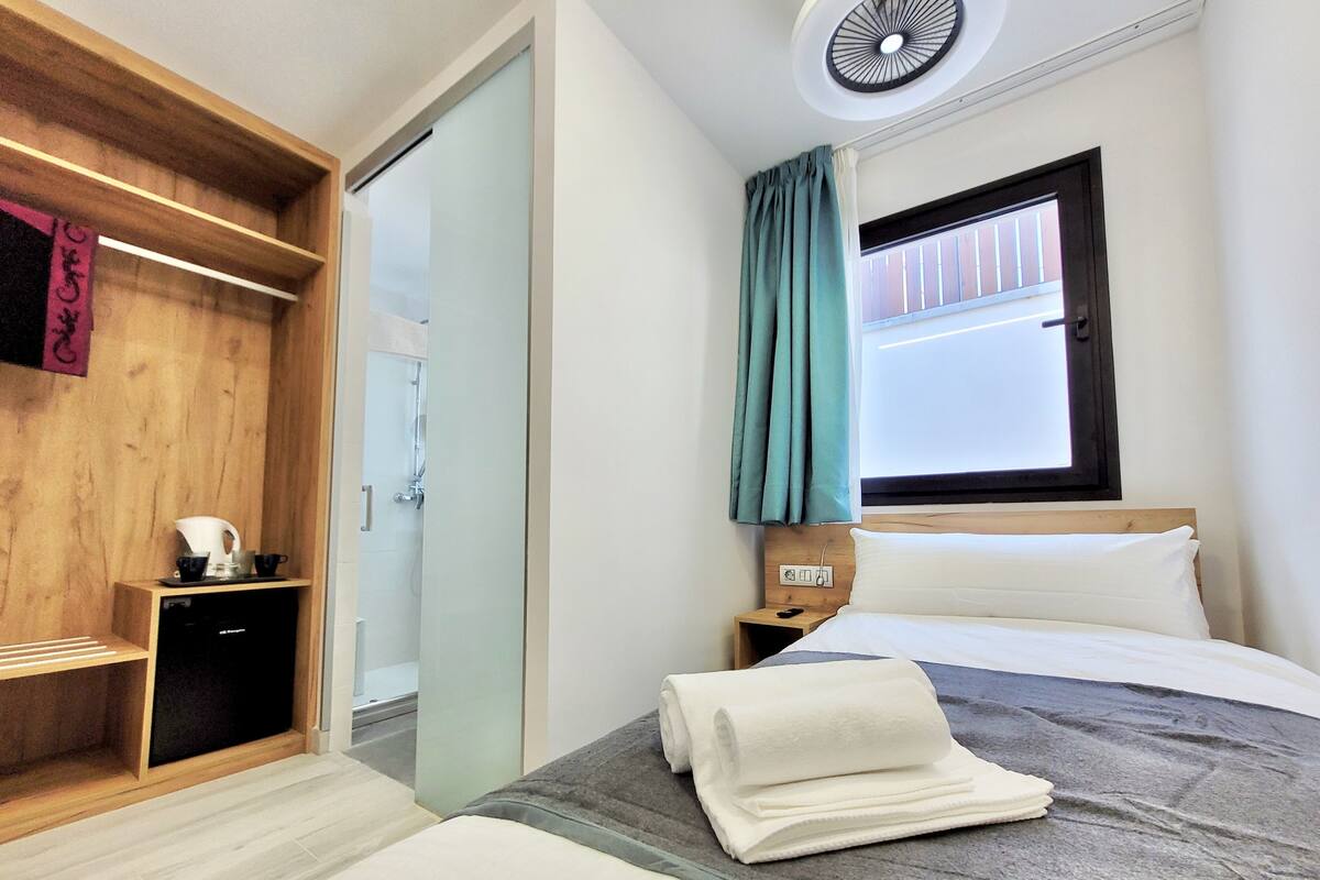 Une chambre d’hôtel comprenant un lit, une table de chevet, une armoire en bois, une salle de bain avec douche et une fenêtre avec des rideaux.