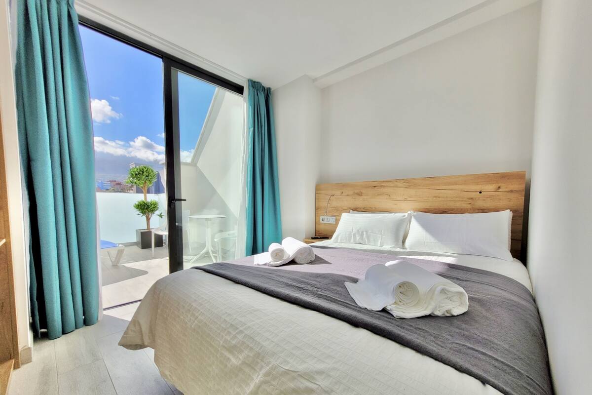 Une chambre d’hôtel avec un grand lit, une tête de lit en bois, des oreillers blancs et une vue sur le ciel par une porte-fenêtre coulissante.