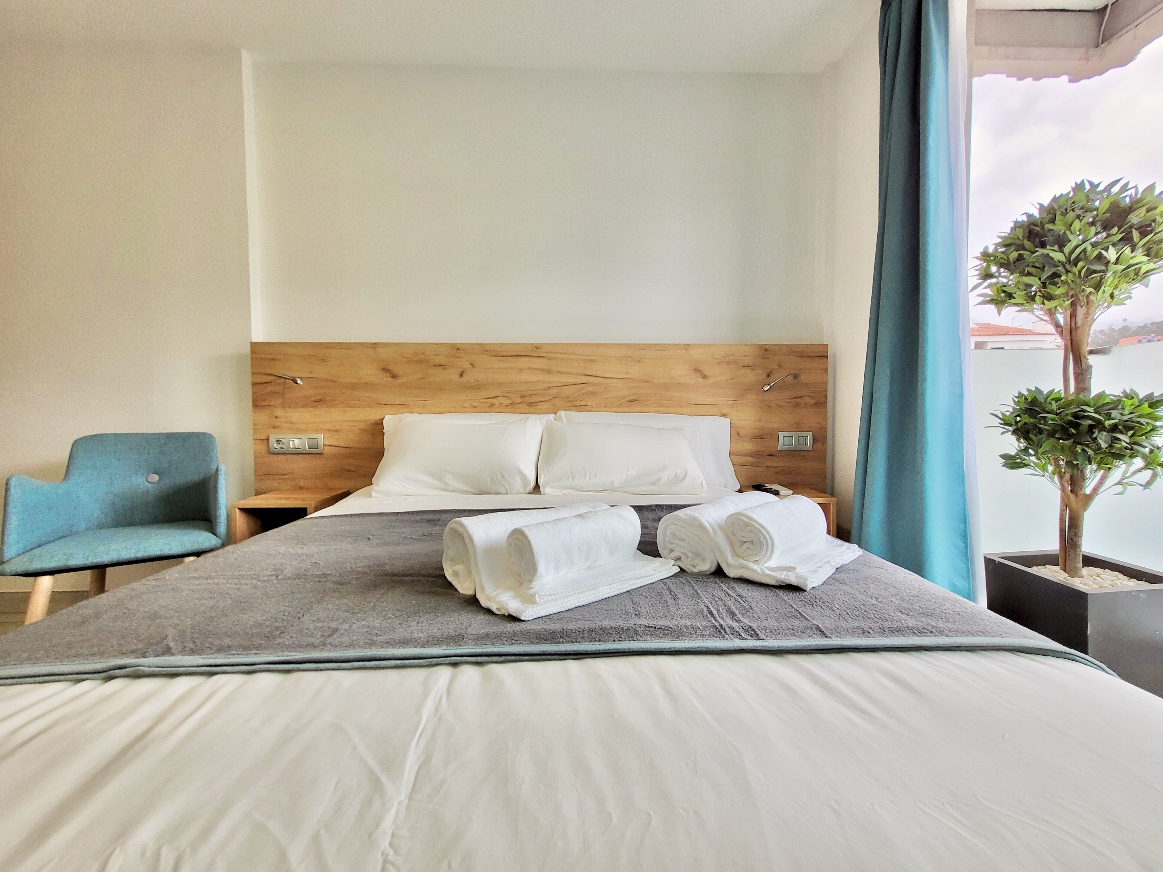 Une chambre d’hôtel avec une tête de lit en bois, un lit avec du linge de lit blanc, une chaise bleue et une plante en pot près de la fenêtre.