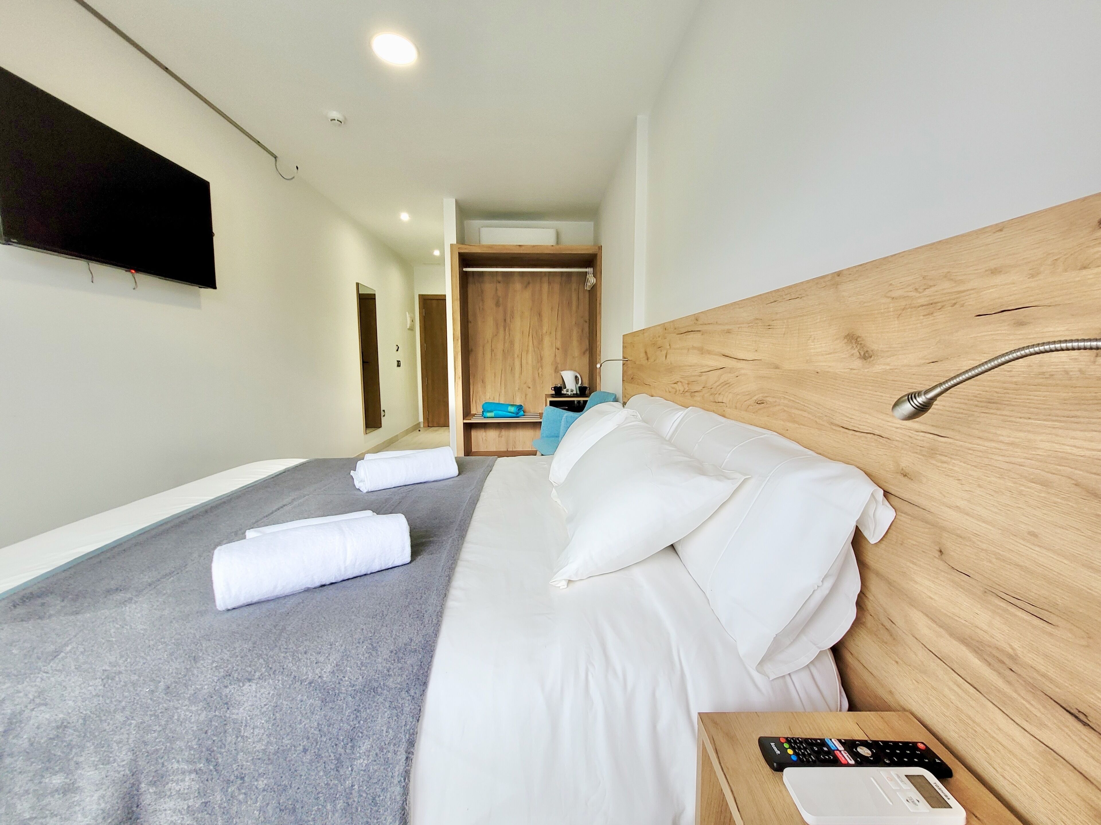 A modern hotel room with a large bed, a wooden headboard, a bedside table with a remote control, and a bathroom visible in the background.