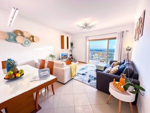 A modern living room with a dining area, a sofa, a TV, and a balcony with a city view.