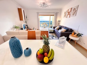 Una sala de estar moderna con comedor, un sofá, una mesa baja con fruta y vista a la ciudad.
