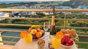 A table with a bottle of champagne, glasses of orange juice, a bowl of fruits, and pastries on a balcony overlooking a scenic view.