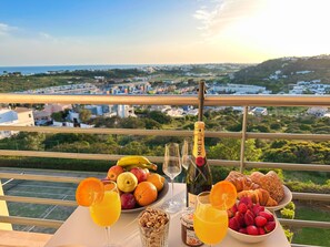 A table with a bottle of champagne, glasses of orange juice, a bowl of fruits, and pastries on a balcony overlooking a scenic view.