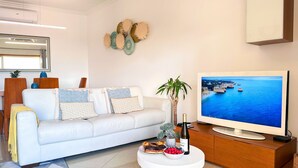 A living room with a white sofa, a television, a coffee table with a bottle and fruit, and a decorative wall piece.