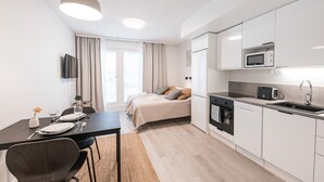 A modern, well-lit room with a dining table set for two, a bed with pillows, a kitchen area with appliances, and a television mounted on the wall.
