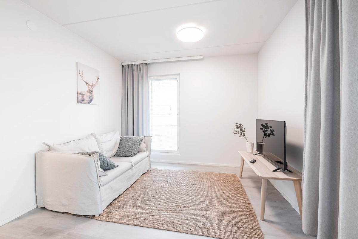 A modern living room with a white sofa, a wooden coffee table, a flat-screen TV, and a large window with curtains.
