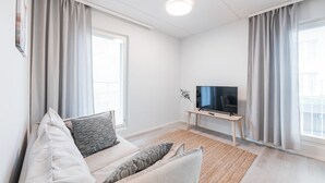 A modern living room with a sofa set, a flat-screen TV on a wooden stand, and large windows with curtains.
