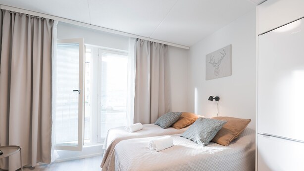 A modern bedroom with two beds, a large window with curtains, a wall-mounted light, and a framed artwork.