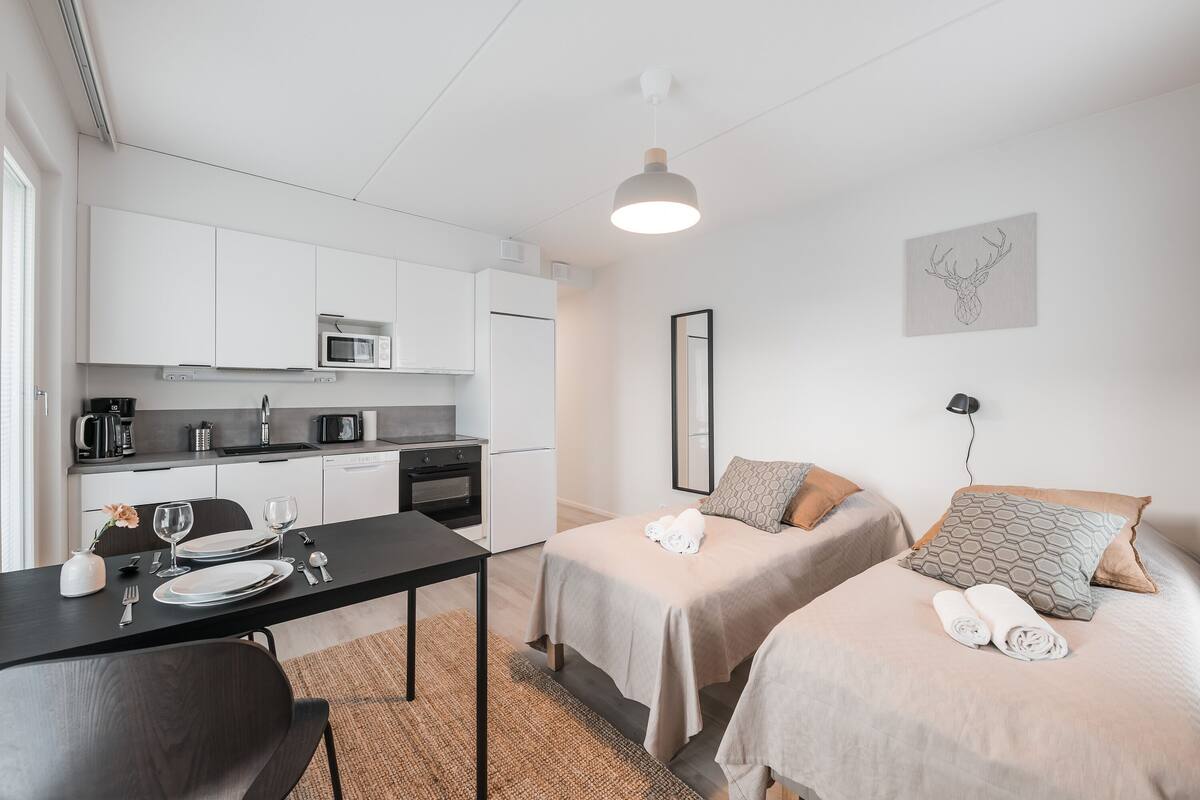 A compact hotel room with a kitchenette, two beds with towels, a dining table, and a wall-mounted artwork.