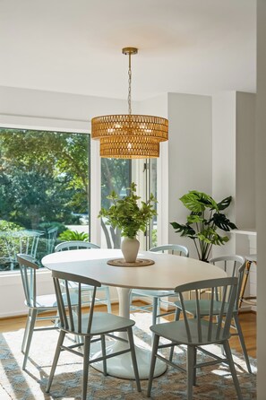 A dining area with a round table, chairs, a hanging light fixture, and potted plants.