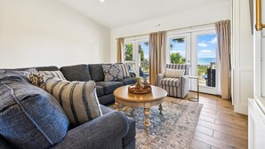 A living room with a dark blue sofa, a round wooden coffee table, and a view of the ocean through large sliding doors. - Welcome to Gulf Dreams! (St. George Island)