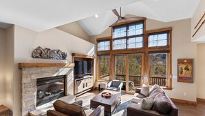A spacious living room with a stone fireplace, comfortable seating, large windows, and a ceiling fan. - Half-Mile to Keystone Base Area! Incredible Slope Views, Private Hot Tub (Keystone)