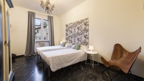 Ett hotellrum med två sängar, en kristallkrona, ett stort abstrakt konstverk på väggen, en brun läderfåtölj och utsikt över byggnader genom ett fönster med gardiner.