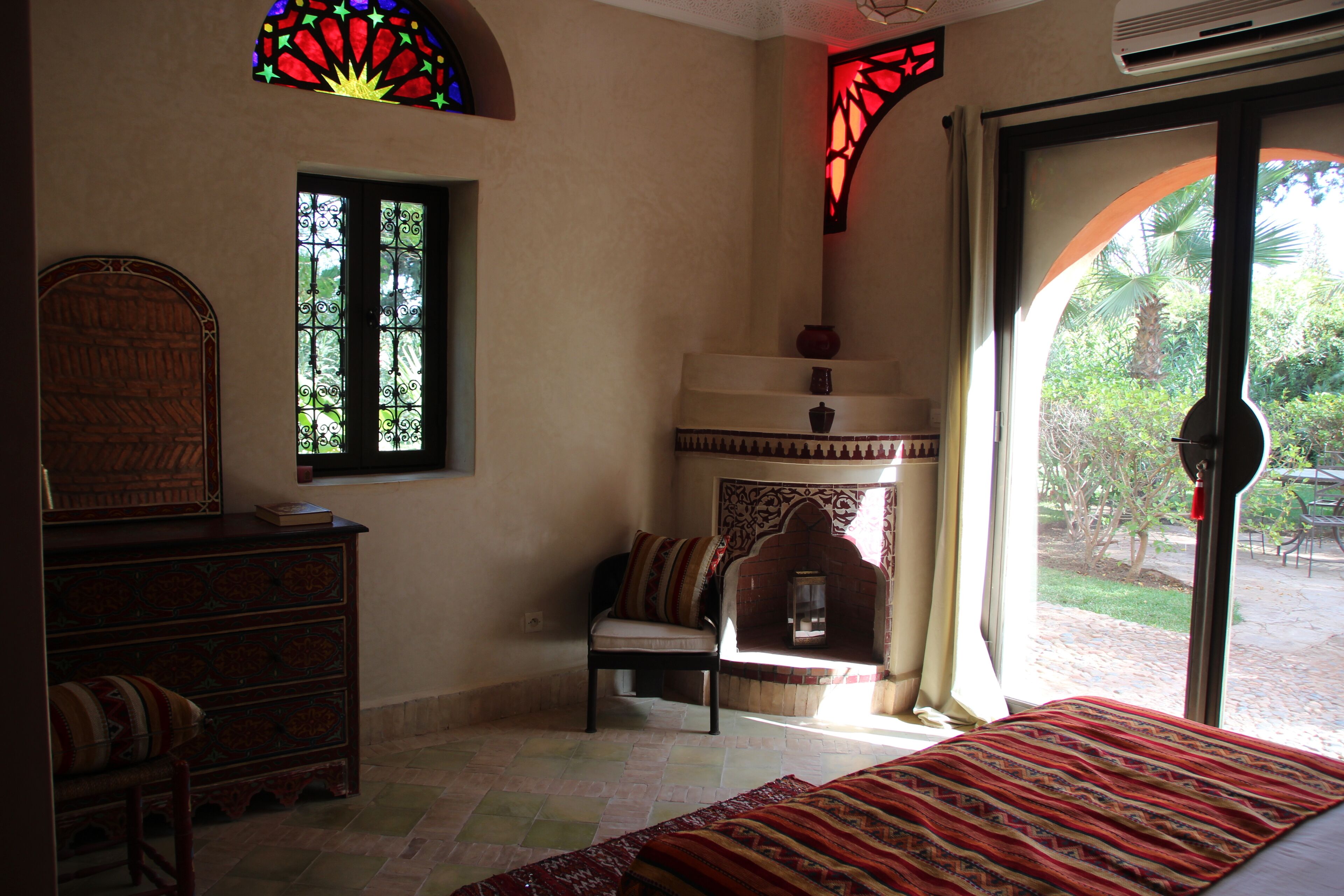A room with a bed, a chair, a wooden cabinet, a window with a decorative frame, and a view of greenery outside.