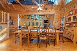 A wooden cabin interior with a dining table, chairs, a ceiling fan, a fireplace, and a television.