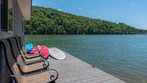 A wooden dock with chairs and a surfboard, overlooking a lake with a forested hill in the background. - Blue Star at Rumbling Bald (Lake Lure)