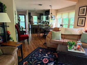 A living room with a sofa, armchair, and a dining table with chairs. There is a kitchen area in the background with a dining table and chairs.