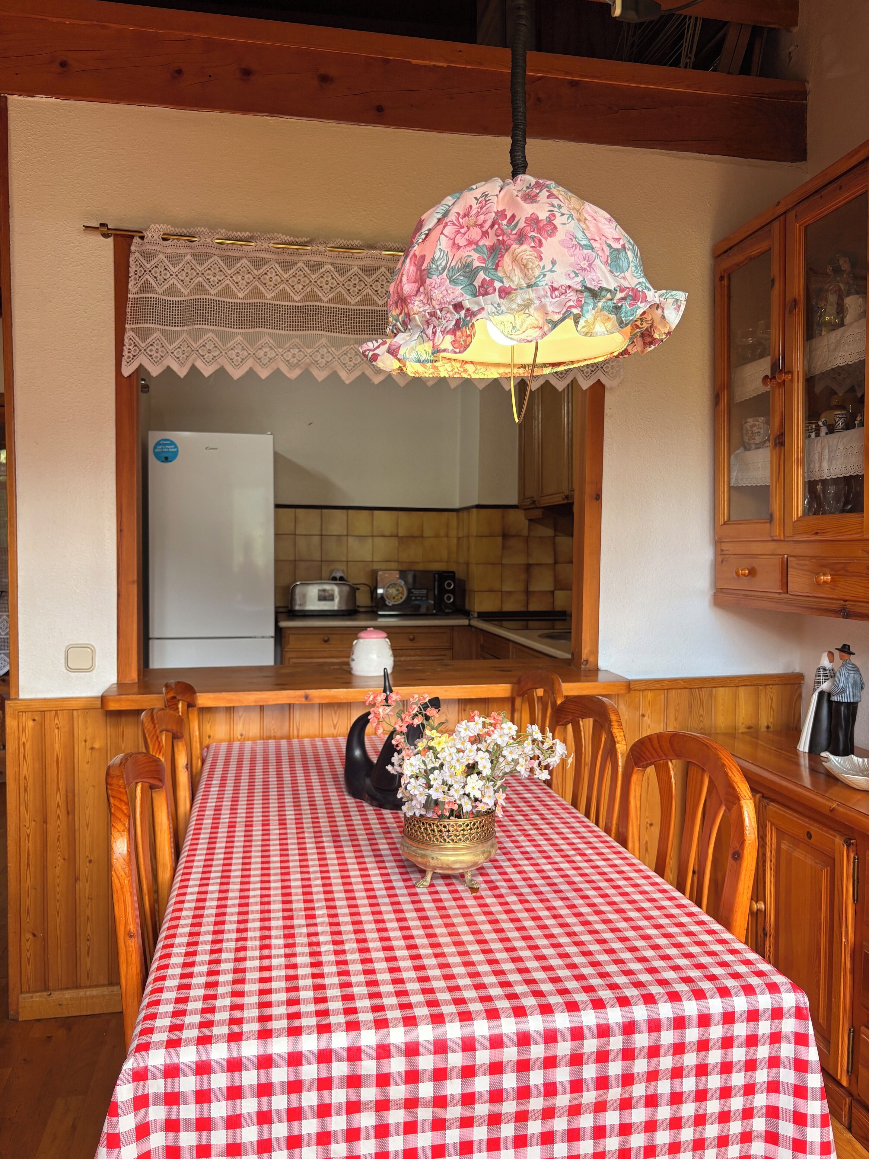 A dining area with a red and white checkered tablecloth, wooden chairs, a floral centerpiece, and a kitchen in the background.
