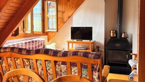 Une pièce mansardée chaleureuse avec un plafond en bois, un téléviseur posé sur un meuble en bois, une cheminée et un coin repas avec une nappe à carreaux.