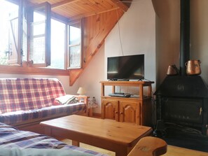 Una habitación acogedora con techo de madera, un televisor sobre un mueble de madera, una chimenea con ollas de cobre, un sofá escocés y una pequeña mesa.