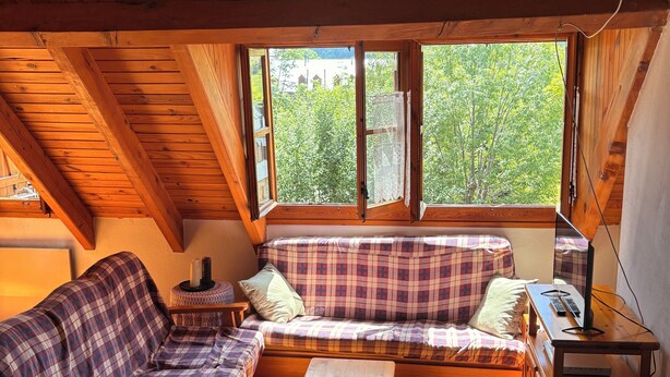 Um quarto de sótão acolhedor com teto de madeira, sofá xadrez, mesa de madeira e vista para o verde lá fora.