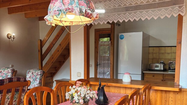 A dining area with a checkered tablecloth, wooden chairs, a floral centerpiece, and a kitchen in the background.
