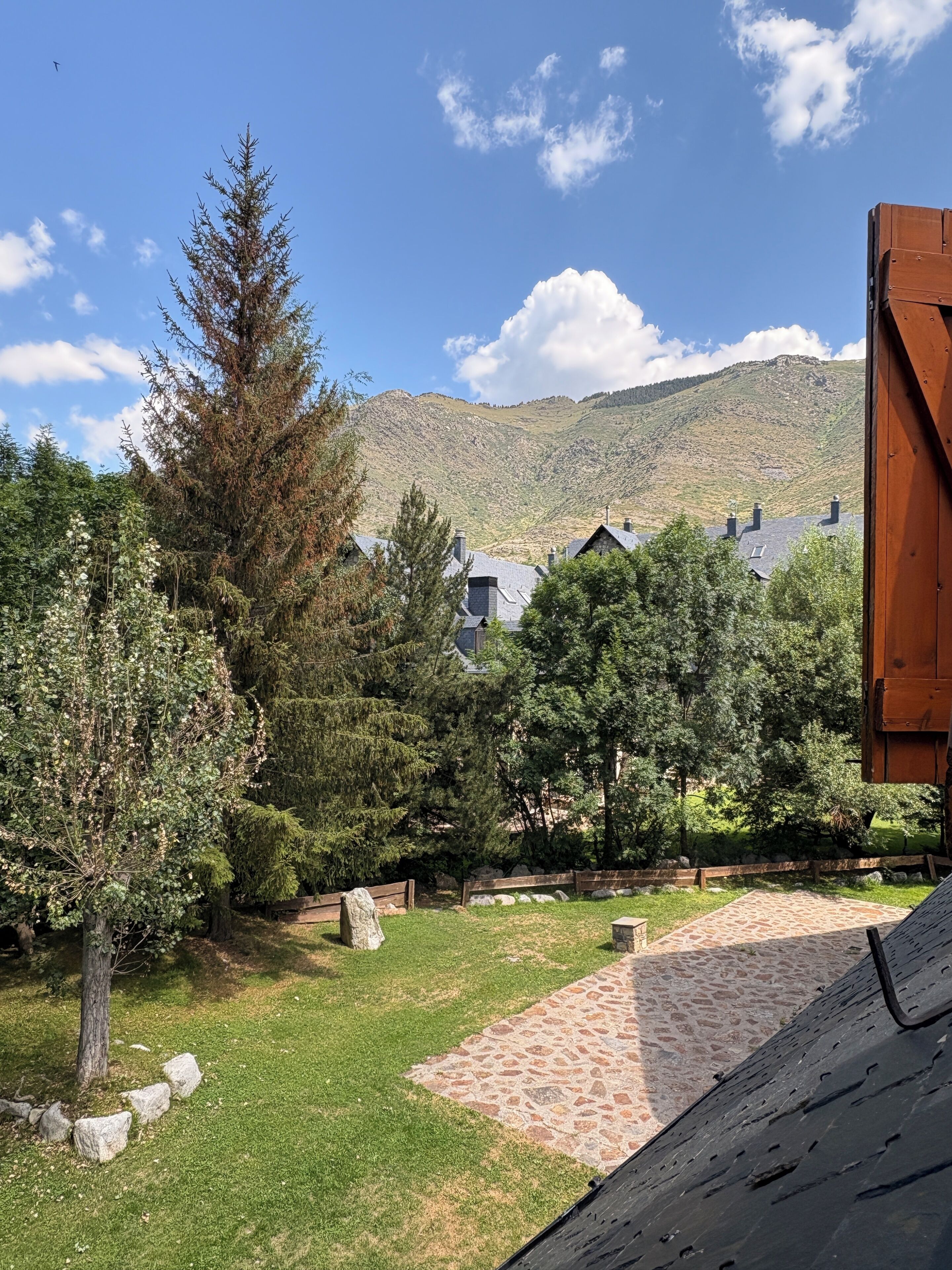 Blick auf eine gebirgige Landschaft mit blauem Himmel, einer Holzhütte auf der rechten Seite und einem Steinweg, der zu einer Sitzgelegenheit führt.