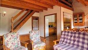 Sala de estar com piso de madeira, dois poltronas florais, um sofá xadrez e uma cozinha visível por uma porta aberta.