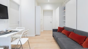 A modern, minimalist living space with a grey sofa, red cushions, a white dining table with chairs, and a flat-screen TV mounted on the wall.