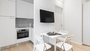 A modern kitchen with white cabinetry, a built-in oven, and a flat-screen TV mounted above a dining table with two white chairs.
