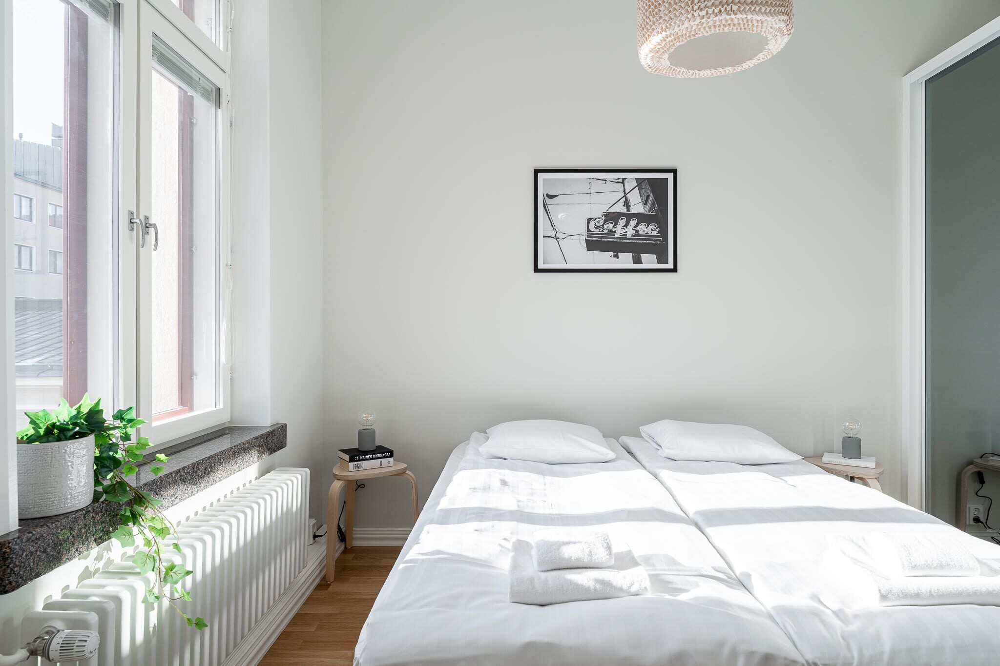 Et soveværelse med seng, radiator, et vindue, en plante, et natbord og et billede på væggen.