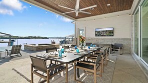 Une terrasse extérieure avec une table dressée pour les repas, des chaises, un ventilateur de plafond et une vue sur une étendue d’eau.