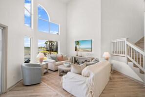 Ein geräumiges Wohnzimmer mit großem Fenster, einer Sofagarnitur, einem Couchtisch und einem Gemälde, das eine Strandansicht zeigt.