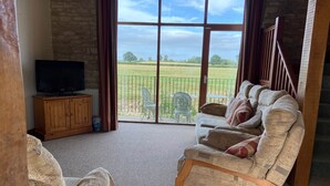 Een woonkamer met een bank, een televisie en uitzicht op een groen veld door grote ramen.