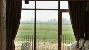 Een kamer met uitzicht op een groen veld door grote ramen, een balkon met een tafel en stoelen, en een bank met gestreepte kussens.